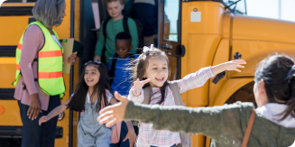 Among a group un-masked school children exiting a school bus, a masked child prepares to embrace a woman