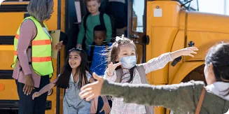 Among a group un-masked school children exiting a school bus, a masked child prepares to embrace a woman